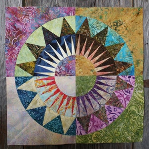 May include: A colourful quilt with a circular design featuring a starburst pattern in the centre. The quilt is made of various fabrics with different patterns and colours, including green, blue, purple, red and yellow.