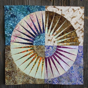 Può includere: Un blocco di trapunta con un design circolare che presenta un motivo a raggiera in tonalità di marrone, viola, blu e bianco. Il tessuto è decorato con motivi floreali e geometrici.