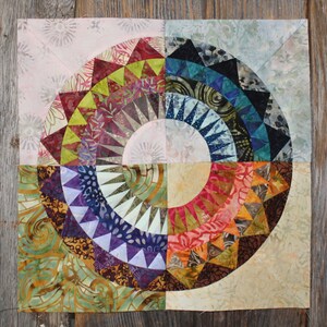Può includere: Un blocco di trapunta colorato con un design circolare che presenta una varietà di tessuti e motivi. Il blocco è composto da 12 sezioni, ciascuna con un design diverso. I colori del blocco includono rosso, arancione, giallo, verde, blu, viola e marrone.