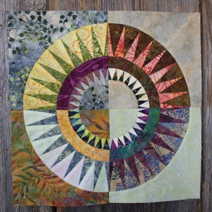 Puede incluir: Un bloque de colcha colorido con un diseño circular que presenta una variedad de telas en tonos de verde, amarillo, morado, rojo, marrón, azul y blanco. El centro del círculo es de color beige claro.