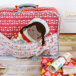 Puede incluir: Una maleta roja y blanca con estampado y ribete azul con una muñeca dentro. La muñeca tiene el pelo castaño, los ojos marrones y un vestido azul y blanco. La maleta tiene un asa en la parte superior y una cremallera.