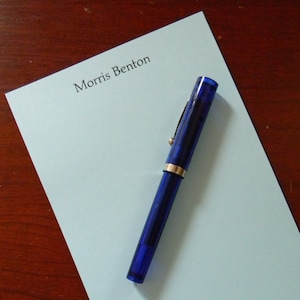 May include: A light blue notepad with the name "Morris Benton" printed at the top. A blue pen is lying on the notepad.