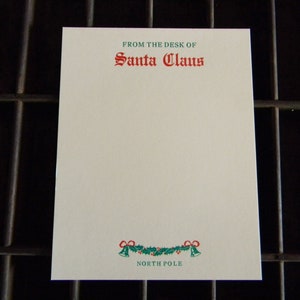 May include: A white notepad with green and red holly and bell border. The text "From the desk of Santa Claus" is printed in green and red at the top of the notepad. The text "North Pole" is printed in green at the bottom of the notepad.
