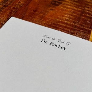 May include: A white piece of paper with the text "From the Desk Of Dr. Rockey" printed in black ink.