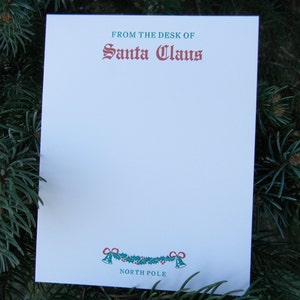 Op de afbeelding: Een witte kaart met groene en rode tekst die "From the desk of Santa Claus" luidt. De kaart heeft onderaan een groene en rode krans met bellen en de tekst "North Pole".