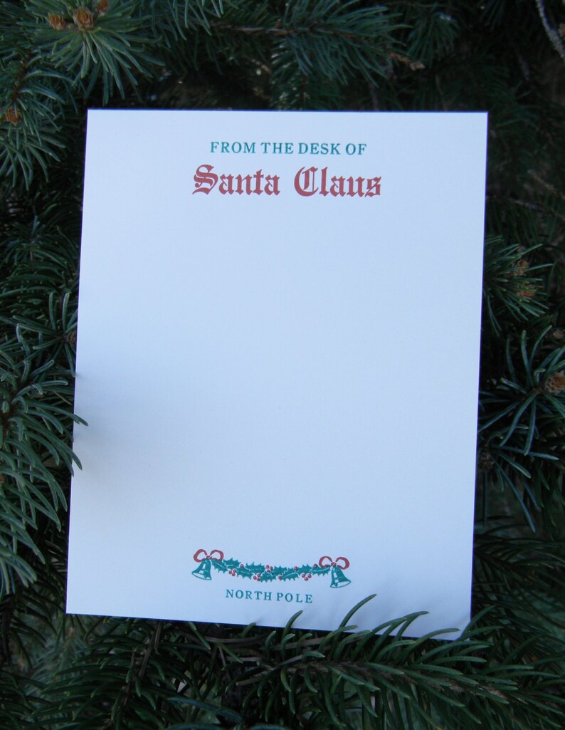 Op de afbeelding: Een witte kaart met groene en rode tekst die "From the desk of Santa Claus" luidt. De kaart heeft onderaan een groene en rode krans met bellen en de tekst "North Pole".