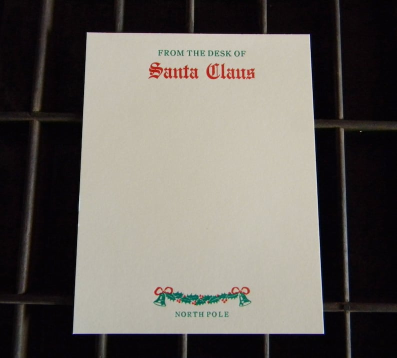 Op de afbeelding: Een wit notitieblok met een groene en rode rand van hulst en bellen. De tekst "From the desk of Santa Claus" staat in groen en rood bovenaan het notitieblok gedrukt. De tekst "North Pole" staat in groen onderaan het notitieblok gedrukt.