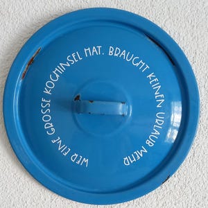 Può includere: Un coperchio rotondo in metallo smaltato blu con una maniglia centrale e il testo stampato in bianco in tedesco: Wer eine große Kochinsel hat, braucht keinen Urlaub mehr. Il coperchio mostra segni di usura con piccole macchie di ruggine sui bordi.