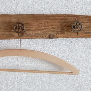 Könnte beinhalten: Ein rustikaler Holz-Kleiderhaken mit drei dekorativen Steuerrad-Haken. Ein natürlicher Holzkleiderbügel hängt an einem der Haken. Der Kleiderhaken ist an einer weißen, strukturierten Wand befestigt.