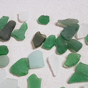 May include: A collection of sea glass in various shades of green and white. The sea glass is arranged on a white fabric background.