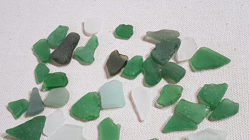 May include: A collection of sea glass in various shades of green and white. The sea glass is arranged on a white fabric background.