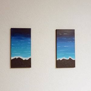 Op de afbeelding: Twee ingelijste schilderijen van zeegezichten met blauw en wit water en een donkerbruin zandstrand aan de onderkant.