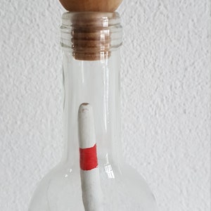 Puede incluir: Una botella de vidrio transparente con un tap&oacute;n de bola de madera y un palo rayado rojo y blanco en su interior.