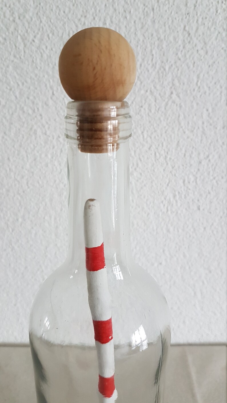 Puede incluir: Una botella de vidrio transparente con un tap&oacute;n de bola de madera y un palo rayado rojo y blanco en su interior.