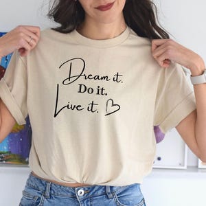Op de afbeelding: Een beige t-shirt met een zwarte afbeelding met de tekst "Dream it. Do it. Live it." met een klein hartje.