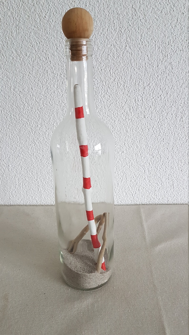 Puede incluir: Una botella de vidrio transparente con un tap&oacute;n de corcho y una tapa de madera. Dentro de la botella hay arena, un palo blanco con rayas rojas y un trozo de madera a la deriva.