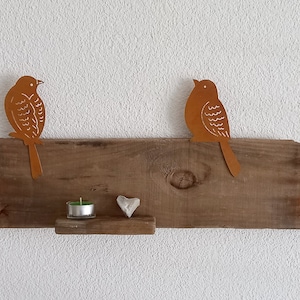 Könnte beinhalten: Ein rustikales Holzregal mit zwei Metallschilden in Form von Vögeln, einem kleinen weißen Herz und einem Teelicht. Das Regal ist an einer weißen Wand montiert.