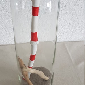 Puede incluir: Una botella de vidrio transparente que contiene un palo rayado de blanco y rojo, arena y peque&ntilde;os trozos de madera a la deriva.