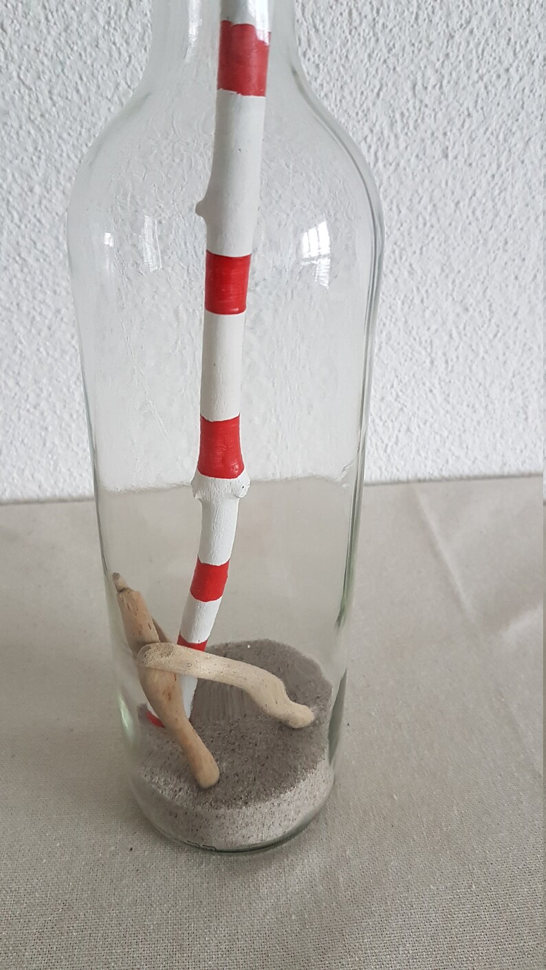 Puede incluir: Una botella de vidrio transparente que contiene un palo rayado de blanco y rojo, arena y peque&ntilde;os trozos de madera a la deriva.