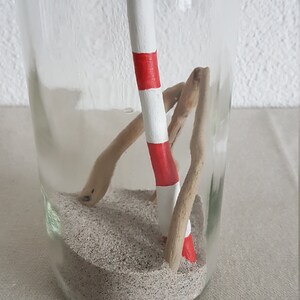 Puede incluir: Un tarro de vidrio con un palo rayado rojo y blanco, un trozo de madera a la deriva y arena en su interior.