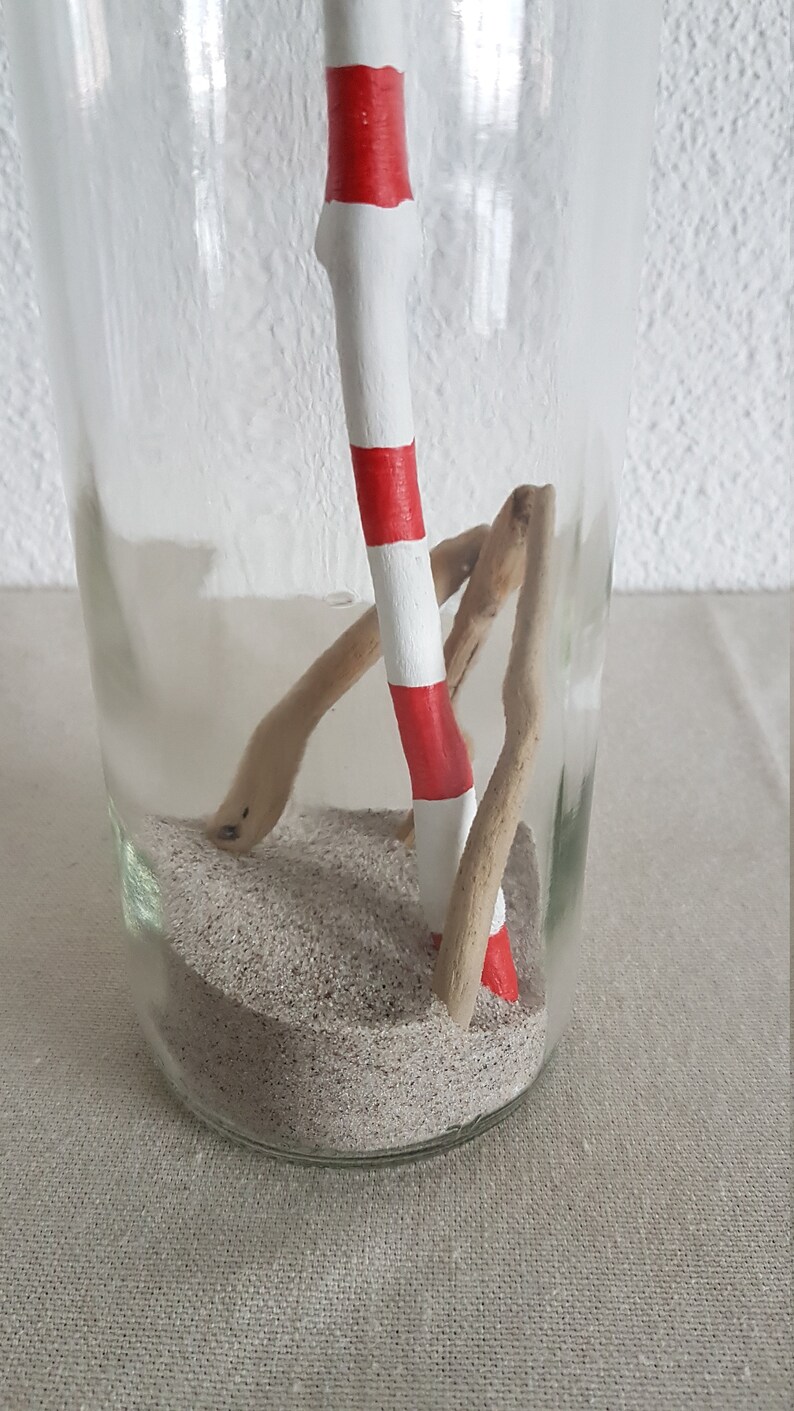 Puede incluir: Un tarro de vidrio con un palo rayado rojo y blanco, un trozo de madera a la deriva y arena en su interior.