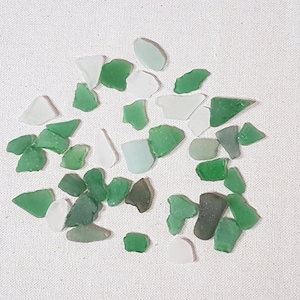 May include: A collection of small, smooth, sea glass pieces in shades of green and white. The glass pieces are scattered on a white fabric background.
