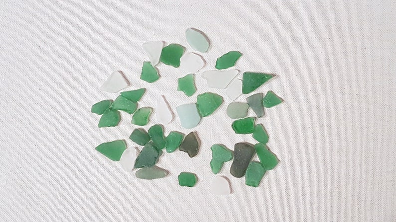 May include: A collection of small, smooth, sea glass pieces in shades of green and white. The glass pieces are scattered on a white fabric background.