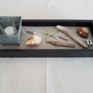May include: A black rectangular tray with a sand-like surface, containing a small metal container with a white candle, a starfish, seashells, and driftwood.