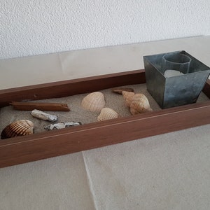 May include: A wooden tray with a light brown finish, filled with sand and seashells. A small metal container with a white interior is placed on the sand.