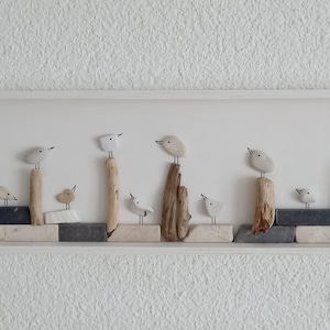 Puede incluir: Una obra de arte mural enmarcada en blanco que presenta una serie de pequeñas esculturas de pájaros de color claro posadas sobre elementos de madera y piedra. Los pájaros están en varias poses, creando una escena serena inspirada en la naturaleza.