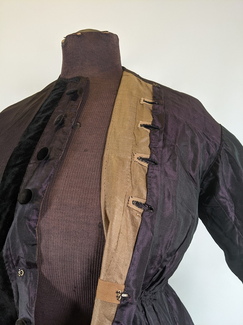 May include: A vintage jacket displayed on a mannequin. The jacket features a dark purple iridescent fabric with black velvet accents and large black buttons. The interior lining is a tan color, and the jacket has a fitted waist.