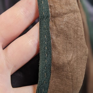 May include: Close-up of a section of fabric with a dark green trim. The fabric is a light brown color and appears to be a woven material. The trim is a tightly woven braid, and the stitching is visible along the edge.