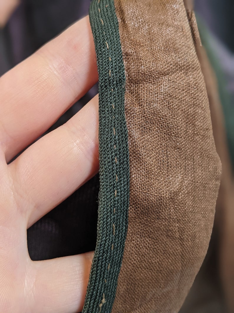 May include: Close-up of a section of fabric with a dark green trim. The fabric is a light brown color and appears to be a woven material. The trim is a tightly woven braid, and the stitching is visible along the edge.
