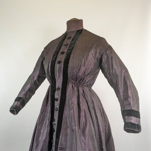 May include: A vintage purple dress with long sleeves and a high collar. The dress features black velvet trim down the front and on the cuffs. It has a fitted bodice and a flared skirt, showcasing a classic silhouette.
