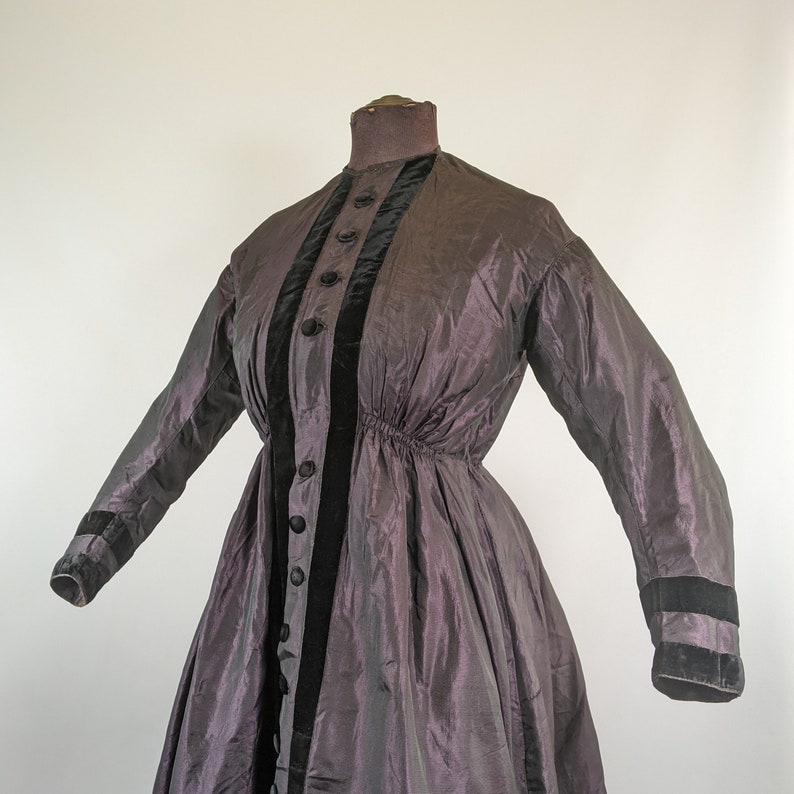 May include: A vintage purple dress with long sleeves and a high collar. The dress features black velvet trim down the front and on the cuffs. It has a fitted bodice and a flared skirt, showcasing a classic silhouette.