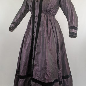 May include: A vintage, floor-length dress in a deep purple hue. The dress features long sleeves, a high collar, and a series of buttons down the front. Black velvet trim accents the front, cuffs, and hem. The dress is displayed on a mannequin.