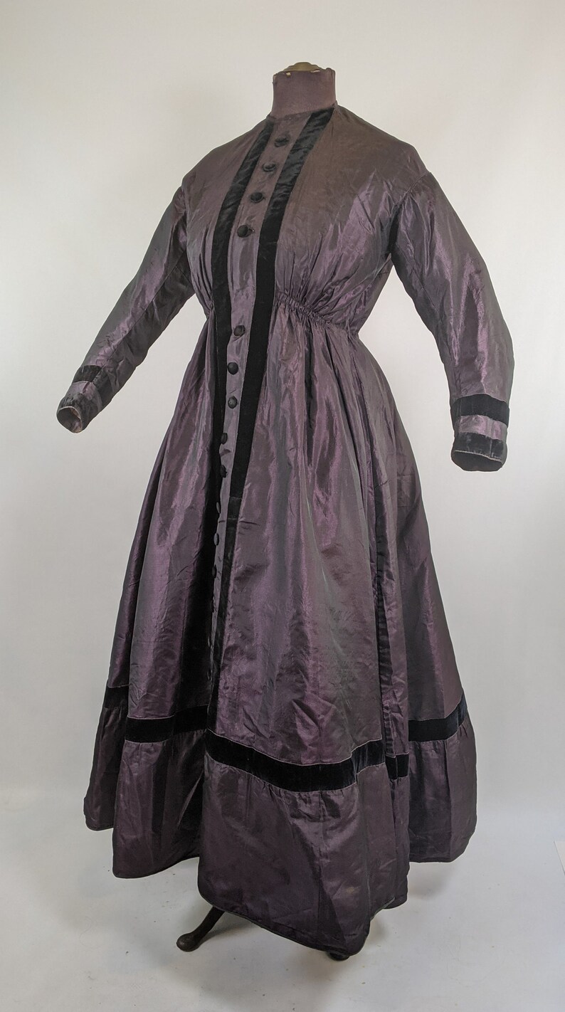 May include: A vintage, floor-length dress in a deep purple hue. The dress features long sleeves, a high collar, and a series of buttons down the front. Black velvet trim accents the front, cuffs, and hem. The dress is displayed on a mannequin.