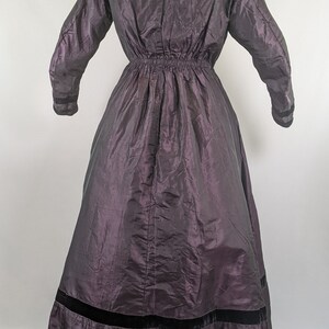 May include: A dark purple vintage dress with long sleeves and a high collar. The dress features a gathered waist and a wide black velvet band at the hem. The fabric has a subtle sheen.