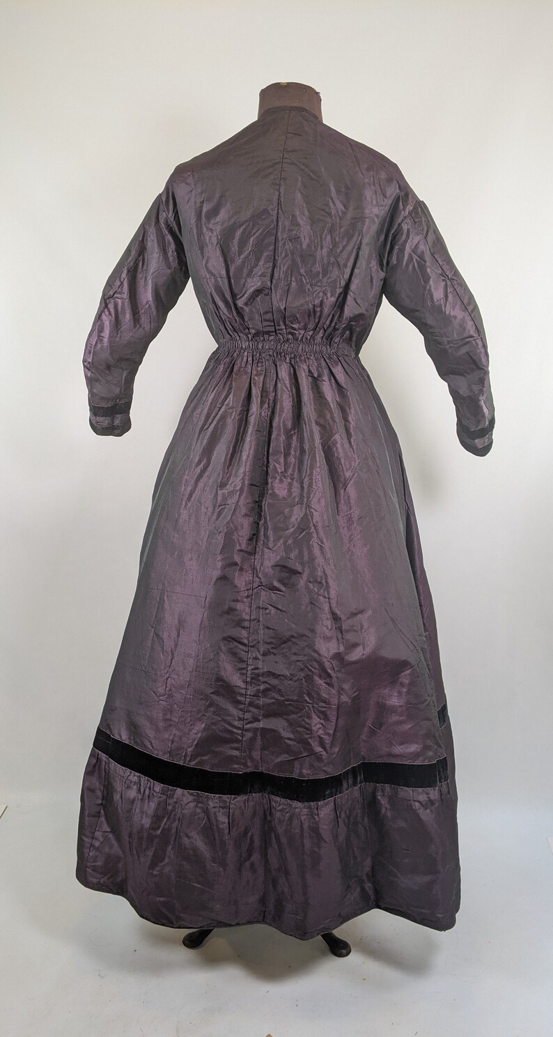 May include: A dark purple vintage dress with long sleeves and a high collar. The dress features a gathered waist and a wide black velvet band at the hem. The fabric has a subtle sheen.