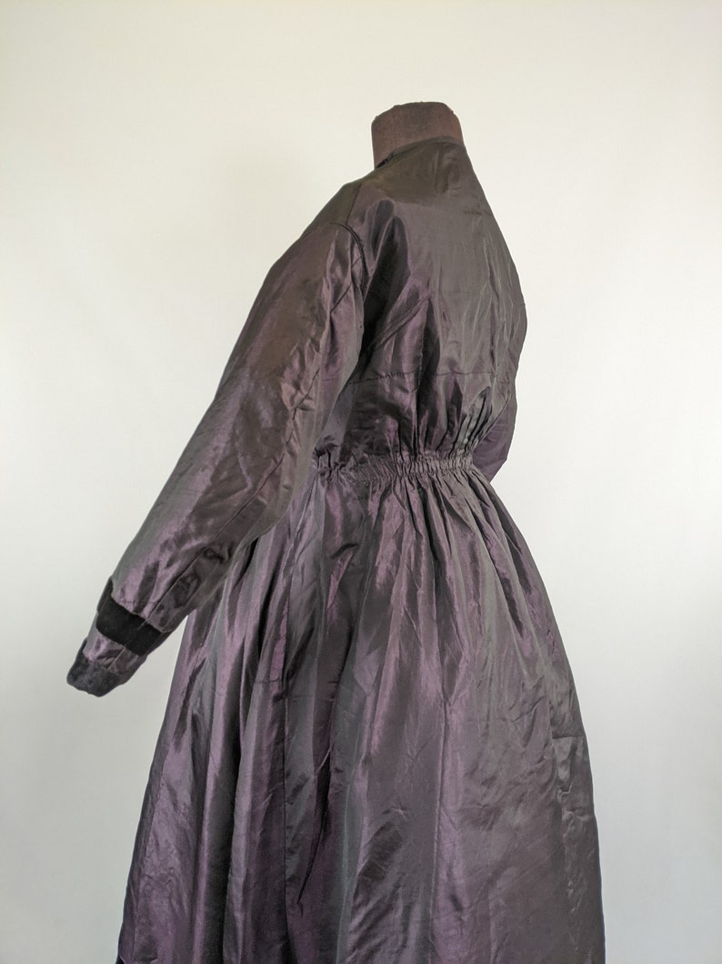 May include: A dark purple vintage coat with a high collar and long sleeves. The coat has a cinched waist and a full skirt. The sleeves have black velvet cuffs. The coat is made of a shiny, iridescent fabric.