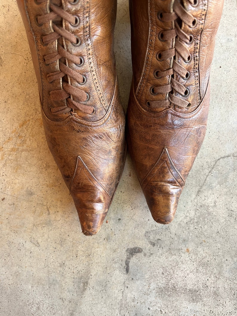 K&ouml;nnte beinhalten: Ein Paar braune Lederstiefel mit Schn&uuml;rung und spitz zulaufenden Zehen. Die Stiefel haben einen abgenutzten Look und sind mit braunen Schn&uuml;rsenkeln gebunden.