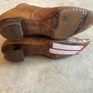 K&ouml;nnte beinhalten: Nahaufnahme der Unterseite von zwei braunen Lederstiefeln mit einem handgeschriebenen Zettel, der an einem Stiefel befestigt ist und lautet: "Jims h&ouml;lzerne, hochhackige Kuhschuhe, die vom Inhaber gekauft wurden, waren 1877 im Trend und kosteten 7,00 $. Charles, ein einzelnes Pferd, war 1877 100,00 $ wert."