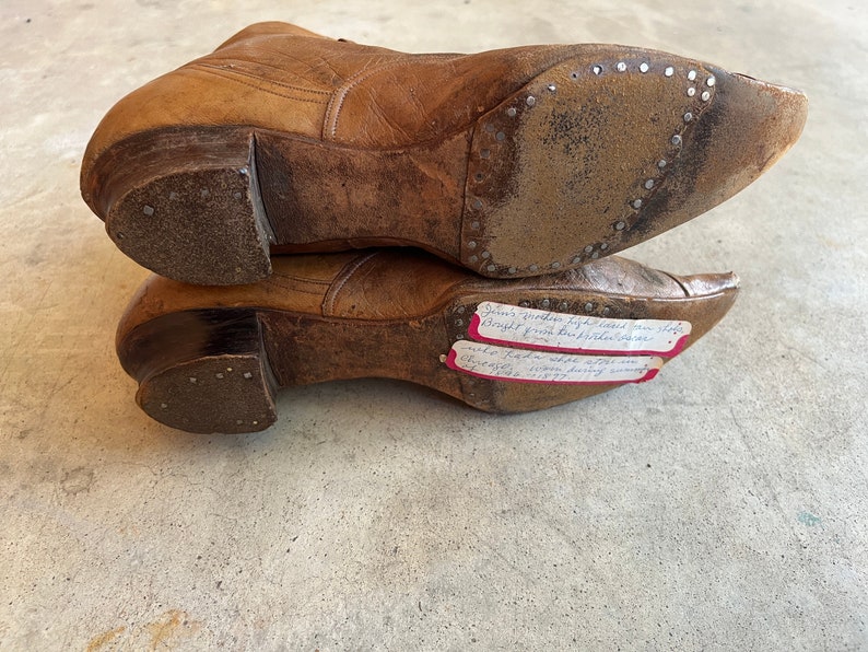 K&ouml;nnte beinhalten: Nahaufnahme der Unterseite von zwei braunen Lederstiefeln mit einem handgeschriebenen Zettel, der an einem Stiefel befestigt ist und lautet: "Jims h&ouml;lzerne, hochhackige Kuhschuhe, die vom Inhaber gekauft wurden, waren 1877 im Trend und kosteten 7,00 $. Charles, ein einzelnes Pferd, war 1877 100,00 $ wert."