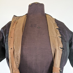 May include: A black jacket with a brown interior and tan trim is displayed on a mannequin. The jacket has a textured appearance and is open, revealing the lining and button closures. The mannequin is a dark brown color.