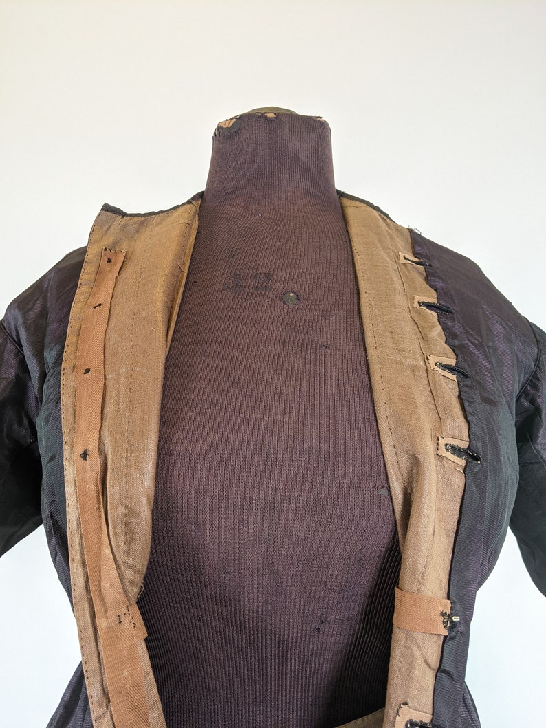 May include: A black jacket with a brown interior and tan trim is displayed on a mannequin. The jacket has a textured appearance and is open, revealing the lining and button closures. The mannequin is a dark brown color.