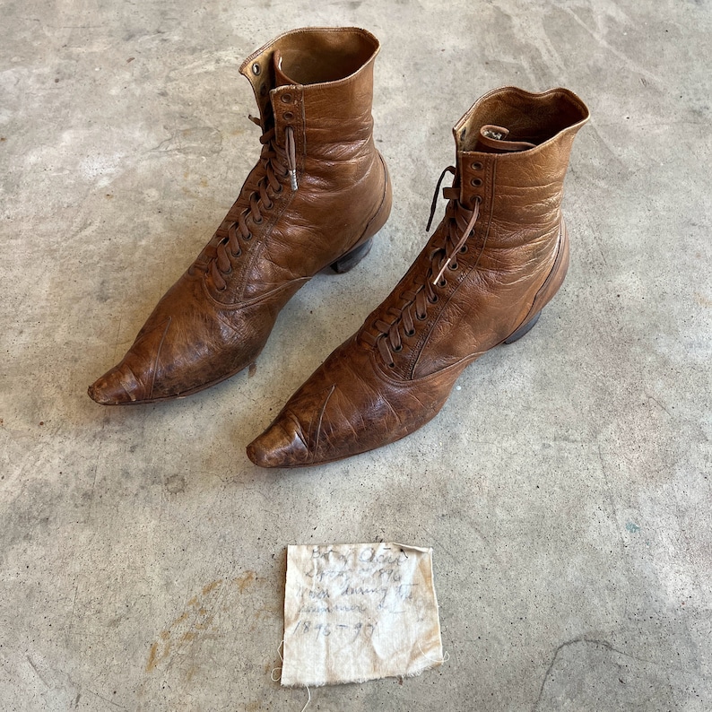 K&ouml;nnte beinhalten: Ein Paar braune Lederstiefel mit spitz zulaufenden Zehen und Schn&uuml;rung. Die Stiefel haben einen Vintage-Look und sind in gutem Zustand. Die Stiefel stehen auf einer Betonfl&auml;che mit einem kleinen St&uuml;ck Papier mit handschriftlichem Text.