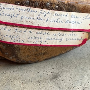 K&ouml;nnte beinhalten: Nahaufnahme eines braunen Lederschuhs mit zwei handgeschriebenen Etiketten. Die Etiketten beschreiben die Schuhe als hochgeschn&uuml;rte braune Schuhe, die 1896-1897 in einem Schuhgesch&auml;ft in Chicago gekauft wurden.