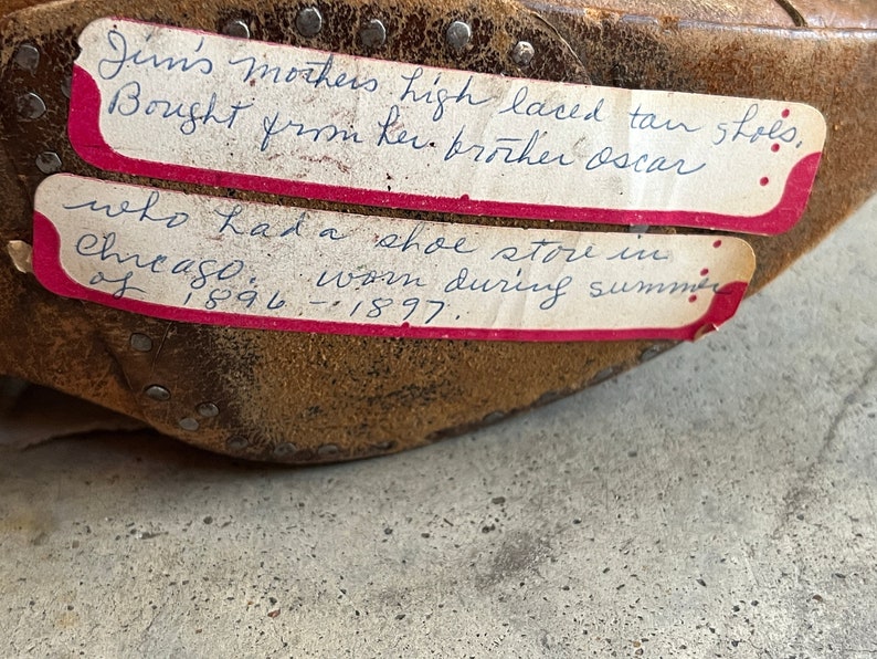 K&ouml;nnte beinhalten: Nahaufnahme eines braunen Lederschuhs mit zwei handgeschriebenen Etiketten. Die Etiketten beschreiben die Schuhe als hochgeschn&uuml;rte braune Schuhe, die 1896-1897 in einem Schuhgesch&auml;ft in Chicago gekauft wurden.