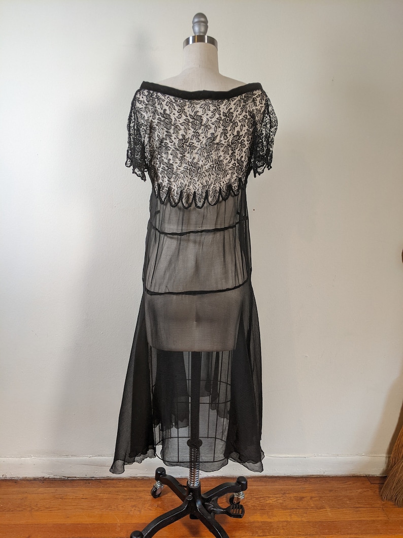 May include: A black sheer dress with a lace bodice and a scalloped hemline. The dress is on a mannequin and has a vintage style.