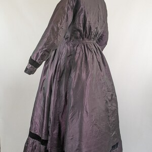 May include: A long, dark purple dress with a high collar and long sleeves. The dress features a gathered waist and a full skirt with black velvet trim at the hem. The fabric has a subtle sheen.